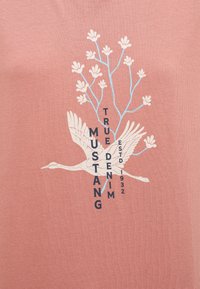 Camiseta de algodón rosa con un gráfico de grulla blanca y un diseño floral azul, con el texto "MUSTANG TRUE DENIM ESTD 1932" en negro.