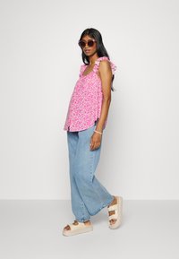 Top sem mangas rosa floral com alças em babado, combinado com jeans de perna larga azul claro e sandálias de plataforma bege. Os acessórios incluem óculos de sol.