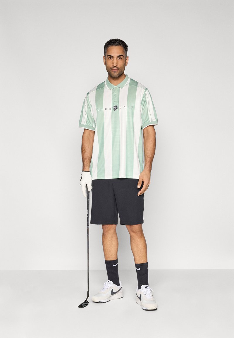 Polo de golf à rayures vert clair et blanc, avec col et logo brodé ; assorti à un short foncé, des baskets blanches et un club de golf.