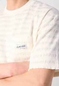 Cremefarvet T-shirt med korte ærmer og horisontale teksturerede striber, med en venstre brystlomme og et synligt brandlogo.