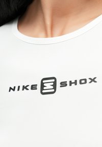 Bijela sportska majica s okruglim izrezom i crnim logotipom s tekstom "NIKE" i "SHOX" te jedinstvenim grafičkim elementom u sredini.