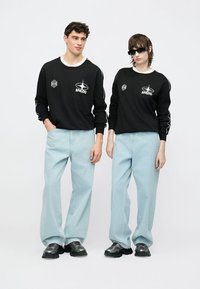 Sweatshirts de algodão pretos com logótipo branco e detalhes em branco, combinados com jeans azul claro de perna larga. Ambos usam ténis grossos pretos e óculos de sol.