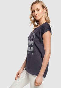 Tricou scurt gri închis cu mânecă scurtă, cu inscripția ��„Bine ai venit în Upside Down” tipărită cu litere mari albe, asortat cu pantaloni albi.