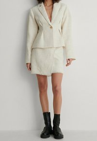 Veste blanche texturée avec jupe mini assortie, présentant des bords effilochés. Portée avec des bottines noires à talons. Coupe lisse et ajustée. Détail de bouton rond.