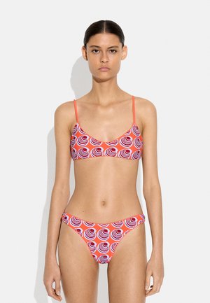 Modelka ubrana w czerwony bikini z białymi i fioletowymi okrągłymi wzorami, stojąca na tle jednolitego jasnego tła.