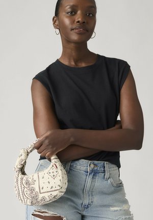 Kvinde med mørk hud iført en ærmeløs sort top og revnede lyseblå jeans, der holder en lille hvid mønstret håndtaske med begge hænder.