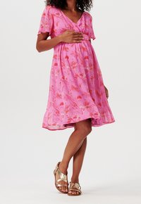 Robe fleurie rose à manches courtes, col en V, froncée sous la poitrine, avec une jupe évasée. Portée avec des sandales à lanières dorées.