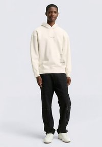 Kremowa bluza z kapturem z kieszenią kangura, detal z logo po boku; zestawiona z czarnymi spodniami cargo z bocznymi kieszeniami, noszona z białymi trampkami.