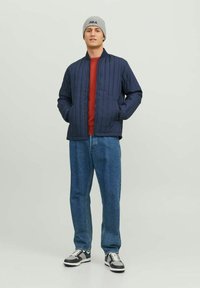 Giubbotto trapuntato blu navy con colletto alto, camicia rossa sotto, jeans blu, berretto lavorato a maglia grigio e sneakers bianco e nero.