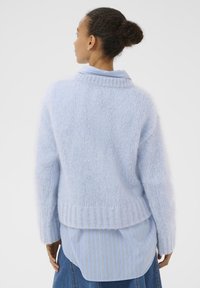 Pull tricoté bleu clair à coupe décontractée, avec ourlet et poignets côtelés, porté par-dessus une chemise à col rayée bleu clair et blanc.