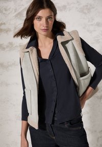 Gilet grigio chiaro senza maniche con foderatura in shearling beige. Caratterizzato da una chiusura con zip e tasche laterali. Indossato sopra una camicia blu navy con bottoni.