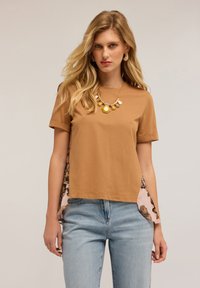 Hnědé bavlněné tričko s krátkým rukávem, které má květinové vzorované boční panely v růžové barvě, zkombinované se světlými modrými džíny.