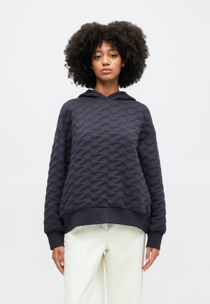 Sweat-shirt à capuche marine avec motif texturé, poignets et ourlet côtelés, présentant une coupe décontractée. Porté avec un pantalon de couleur claire.