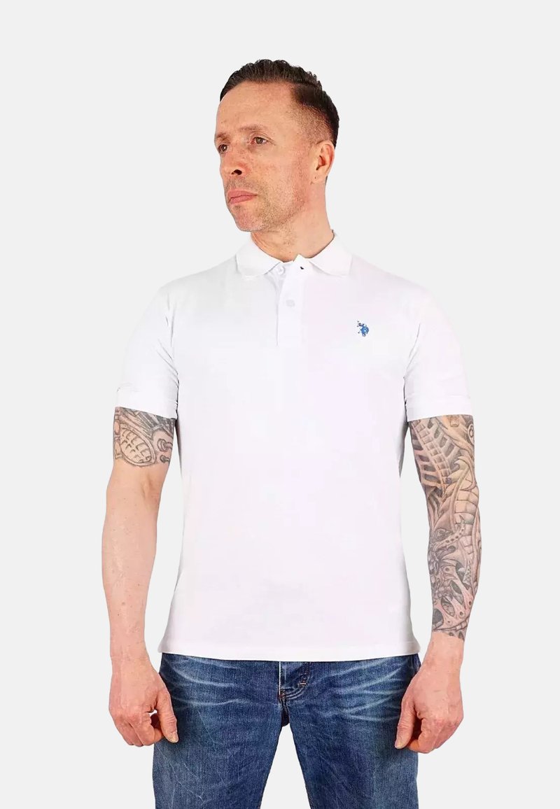 Polo shirt bianco a maniche corte con colletto e logo ricamato blu sul lato sinistro del petto, indossato con jeans in denim blu.