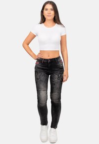 Jeans neri skinny con texture strappata, multiple tasche e cuciture di contrasto rosse, abbinati a un crop top bianco aderente e sneakers bianche.