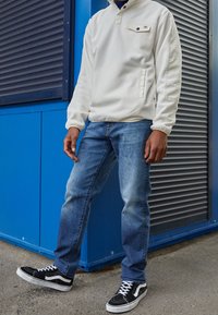 Sudadera de forro polar blanca con un collar de media botonadura, bolsillo delantero, jeans azules y zapatillas Vans negras con suelas y cordones blancos.