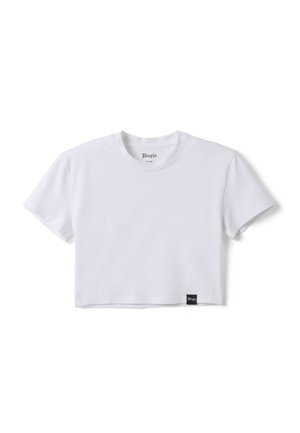 Junge Frau mit langen dunklen Haaren, die ein weißes kurzes T-Shirt, lockere weiße Hose und schwarze Flip-Flops trägt, vor einem einfarbigen Hintergrund stehend.