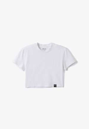 Giovane donna con lunghi capelli scuri che indossa una t-shirt bianca corta, pantaloni bianchi larghi e infradito nere, in piedi davanti a uno sfondo semplice.