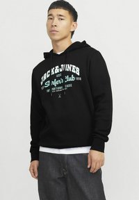 Sudadera con capucha negra que presenta texto en blanco y turquesa que dice "JACK & JONES Surfer's Club 1990" con un corte relajado y un bolsillo tipo canguro en la parte frontal.