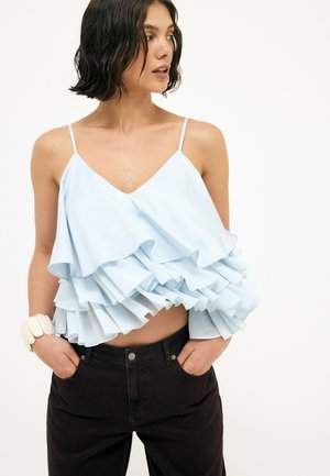 Donna con capelli corti neri che indossa una canotta azzurro chiaro con volant e pantaloni neri, mani nelle tasche, guarda di lato.