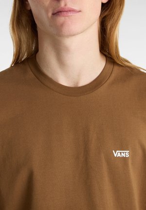 Personne aux cheveux longs portant un t-shirt Vans marron avec un petit logo Vans blanc sur le côté gauche de la poitrine, recadré juste en dessous du nez.