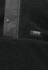 Μαύρος fleece ύφασμα με υφή, που διαθέτει μια λεία μαύρη πάνελ με κουμπί σπασίματος και ετικέτα "TAION Mountain Wear."
