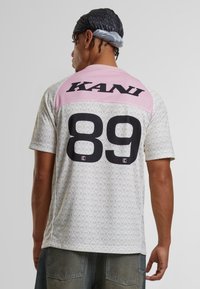 Tričko s krátkým rukávem na bílem vzorovaném podkladu, s růžovými akcenty na ramenou a velkou černou typografií "KANI" a "89" na zadní straně.