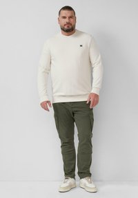 Crèmekleurige trui, olijfgroene cargobroek en witte sneakers. De trui heeft een ronde hals en een klein logo op de borst. Casual en gestructureerde pasvorm.
