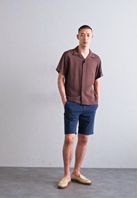 Chemise marron à manches courtes avec une texture douce, shorts navy et mocassins de couleur claire. Design simple avec une coupe détendue et des détails minimalistes.