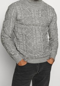 Pull en maille torsadée gris avec un col montant, présentant des motifs texturés et des poignets côtelés. Porté avec un jean foncé, mains dans les poches.