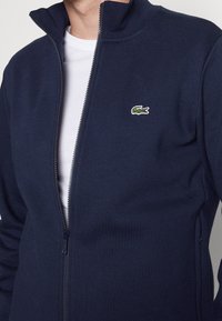 Giacca in pile blu navy con zip e colletto alto, caratterizzata da un piccolo logo ricamato a forma di coccodrillo verde sul lato sinistro del petto. Tessuto morbido, vestibilità regolare.