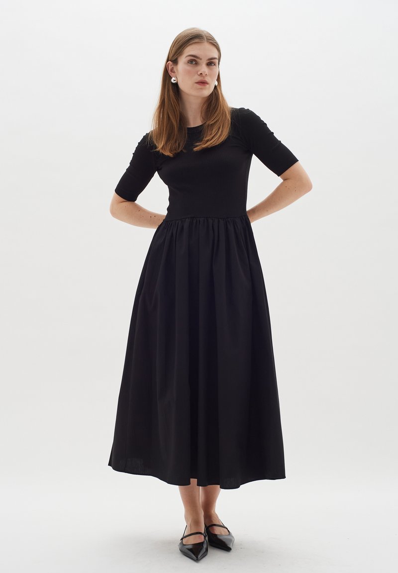 Schwarzes Midi-Kleid mit geripptem Oberteil und ausgestelltem Rock. Kurzärmel und geraffte Taillennaht. Das Modell trägt spitze Ballerinas und runde Ohrringe.