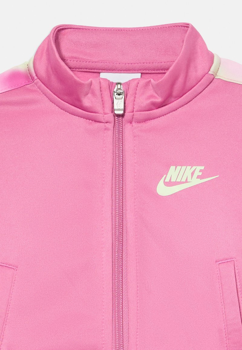 Giacca da track Nike rosa con colletto alto, zip completa e tasche laterali. Presenta un logo Nike bianco sul lato sinistro del petto. Tessuto morbido.