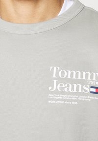 T-shirt gris clair à col rond avec logo blanc "Tommy Jeans", petit drapeau rouge, blanc et bleu, et noms de villes imprimés sur la poitrine.