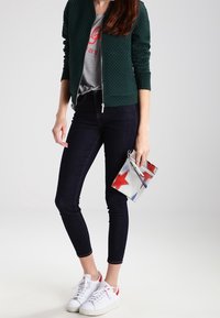 Grön quiltad bomberjacka över en grå grafisk t-shirt, mörka slim fit jeans och vita sneakers med röda accenter, hållande en silverfärgad clutch med stjärnor.