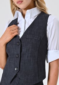 Gilet grigio scuro in tessuto testurizzato con scollatura a V profonda, tre bottoni e una tasca sul petto, sovrapposto a una camicia bianca con colletto.