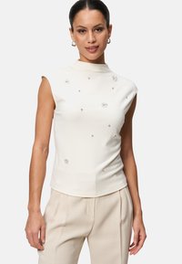 Haut sans manches blanc avec un col montant. Présente des motifs floraux embellis et de petits strass. Texture douce et lisse avec une silhouette ajustée.
