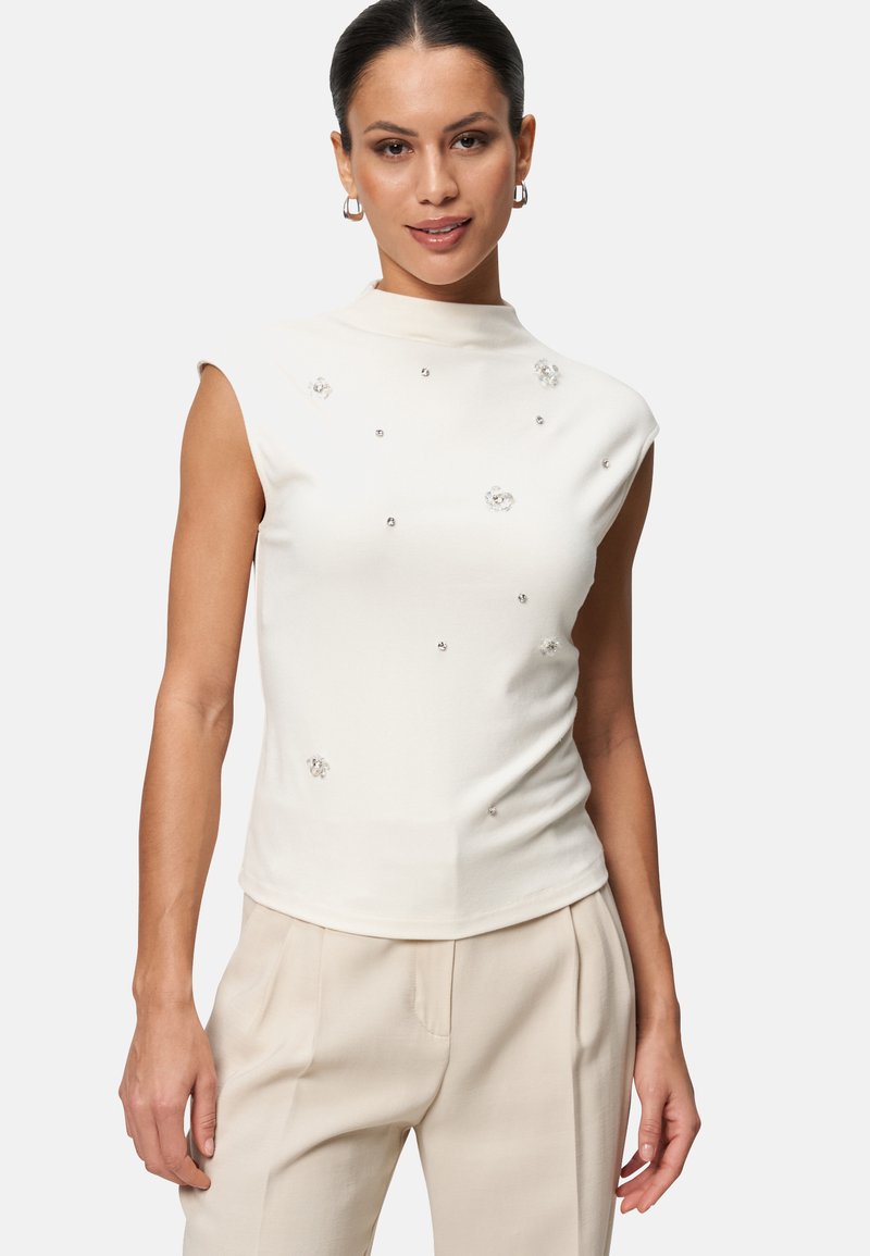Haut sans manches blanc avec un col montant. Présente des motifs floraux embellis et de petits strass. Texture douce et lisse avec une silhouette ajustée.