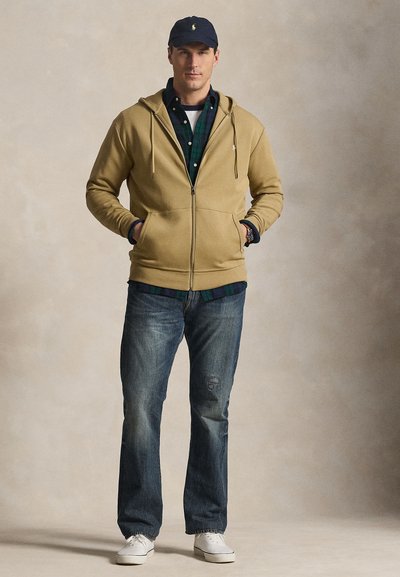 Homme portant un sweat à capuche beige zippé, une chemise à carreaux verte, un jean bleu, des baskets blanches et une casquette bleu marine, debout les mains dans les poches.