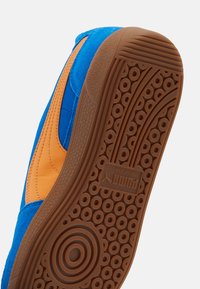 Blå och orange sneakers med övre i mocka, gummisula med cirkulära mönster, och en texturerad, brun yttersula. Puma-logotyp synlig.