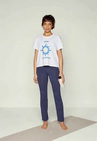 Noor inimene seisab paljajalu jooga matil, hoides vett täis pudelit, kandes valget graafilist T-särki ja tumehallisid pükse, ilus taust on ühtlase värviga.