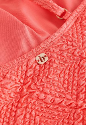 Korallenrosa Kleidungsstück aus textured Stoff, mit Rüschendetails. Ein metallisches Etikett mit einem stilisierten Buchstaben "B" ist auffällig angebracht.