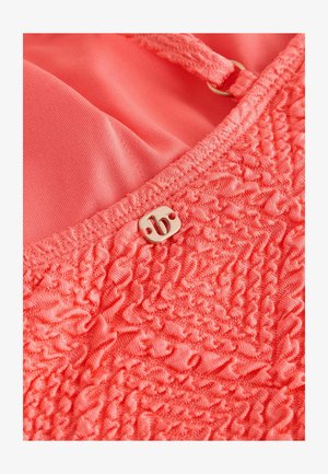 Vêtement rose corail en tissu texturé, présentant des détails froncés. Une étiquette métallique avec une lettre stylisée "B" est fixée de manière proéminente.