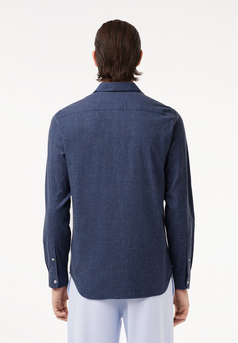 Camicia blu scuro realizzata in un tessuto testurizzato, con maniche lunghe, colletto a punta e dettagli con bottoni sui polsini. L'orlo è leggermente più lungo sul retro.