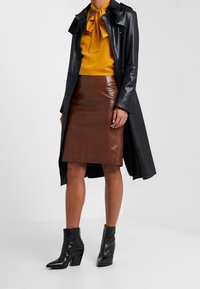 Trench coat in pelle nera, blusa gialla con fiocco, gonna a matita in pelle marrone e stivaletti neri con tacco largo.