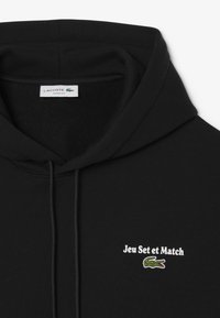 Sudadera negra con cuerda de ajuste, texto "Jeu Set et Match" y logo bordado del cocodrilo verde de Lacoste en el pecho, etiqueta que dice "Lacoste Loose Fit".