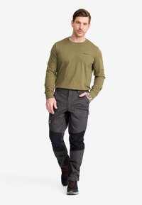 Man som bär en olivgrön långärmad skjorta, mörkgrå cargobyxor med svarta lappar och svarta sneakers med röda snören, står med handen i fickan.