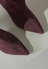 Bordové semišové lodičky so špicatou špičkou, hladkým povrchom a minimálnym šitím. Tiene vrhnuté na štruktúrovaný svetlošedý povrch.