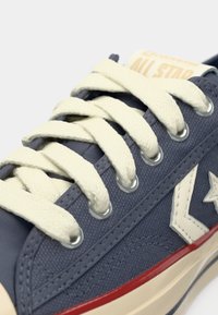 Gros plan d'une basket en toile bleu marine avec un logo étoile blanc, des lacets crème, des œillets métalliques, et un bord de semelle en caoutchouc rouge et crème.