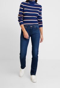 Jersey de cuello alto a rayas azules con rayas naranjas y blancas, combinado con jeans de mezclilla oscuros de corte slim y zapatillas blancas. Diseño sencillo, look casual.